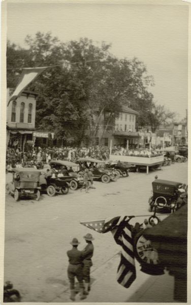 Elevated view of a crowd of people gathering across the street, near a stage. Automobiles are parked in the center of the street near the stage. In the foreground two uniformed soldiers are watching the crowd. Caption reads: "Concert by one of the bands of the 161st Artillery Brigade on the main street of Kilbourn Wis. May 24th 1918. The Brigade laid over at Kilbourn about 36 hours marching out at midnight — May 24."