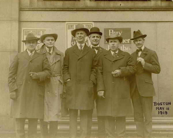 Six men are posing in front a wall where Home Service Fund posters are hanging. Two of the men are smoking cigars. Caption reads: "Delegation to meet 32nd Division after World War I; E.S. Hall, mayor of Green Bay, tallest; Judge Henry Graass at his left and behind him."