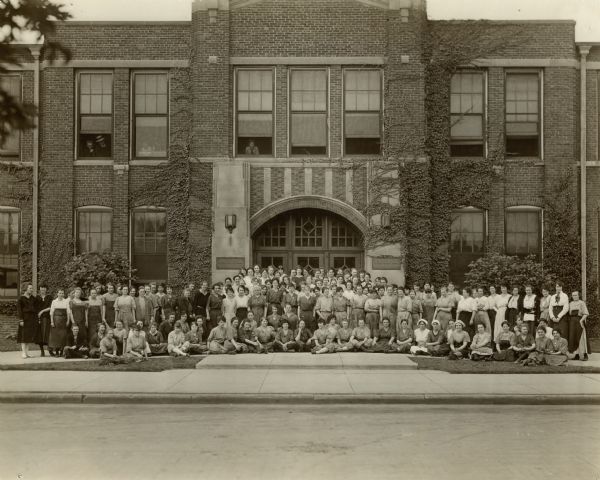 View across street towards a large group of women at the Nash Motor Company building. A few people are looking out from open windows in the building. Caption reads: "Women employed by Nash Motor Company — Kenosha."