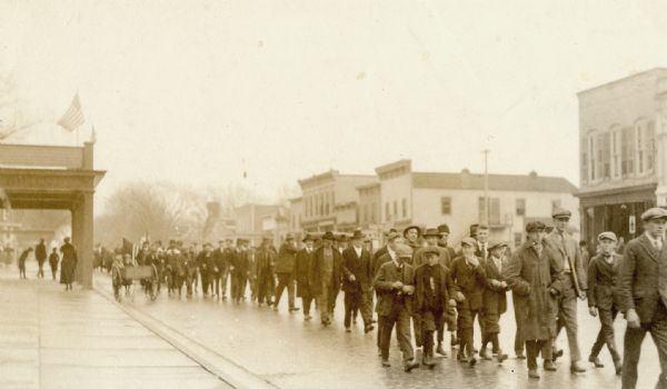 View from sidewalk towards a parade of mostly men and boys walking down a street. A few people are carrying American flags. Caption reads: "Kilbourn, Nov. 11/18. Celebrating Armistice."