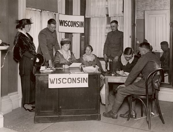 Four women and three men standing or sitting near a desk and table that are labelled "Wisconsin." Two of the women appear to be doing paperwork, and the men are wearing soldier's uniforms. Caption reads: "Wisconsin Hospitality Committee, 1919. New York, N.Y."