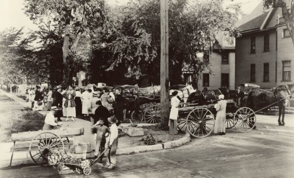 People stand near several carts, which are hitched to horses. In some carts boxes of vegetables are visible. Caption reads: "Madison, Wis. August, 1917. Curb market established by a committee of women of the Dane County Council of Defense Food Board. Opened again in June, 1918. Operated every Tuesday and Thursday from 7-9 AM."