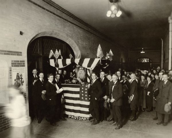 Several young men stand around a kiosk decorated with American flags. One is dressed as Uncle Sam. Behind, them, young men and women are standing. A War Savings Stamps poster is hanging on the wall. Caption reads: "High school boys conducting Liberty Loan Drive, Apr. 1918, Madison, Wis."