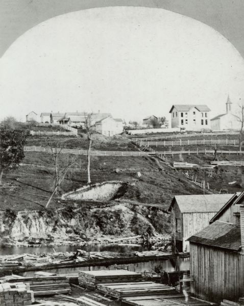 Elevated view of several industrial buildings near water, and in the background are fences on a slope, and at the top of the hill are buildings and what may be a church. In the foreground are stacks of lumber.