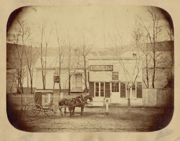 Exterior view of a small bank. A horse-and-carriage is hitched outside the bank, and a house is behind it.