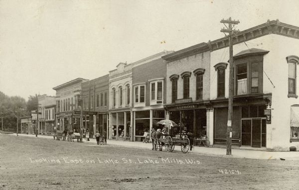 Street view of Lake Street in the town of Lake Mills. A horse-drawn cart has a parasol advertising O&E Patton's Sun-Proof Paints. Businesses in the background include the E.G. Saecker hardware store, the Modern Meat Market, and a lunch and ice cream parlor. Caption reads: "Looking East on Lake St., Lake Mills, Wis."