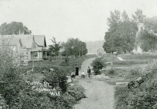 Elevated view of an unpaved street or path in Cazenovia. Three men and a woman are standing and sitting on rocks along the path. Houses and buildings are farther down the road on either side.