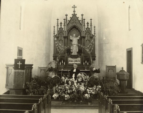 Interior of church, towards altar, pulpit, baptismal font, and pews. Rev. E.F. Scherbel is standing surrounded by baskets of flowers and a banner that reads 50 in honor of the 50th anniversary of his ordination.