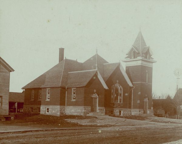 Exterior of a church. Buildings, possibly homes, as well as a windmill are in the background.