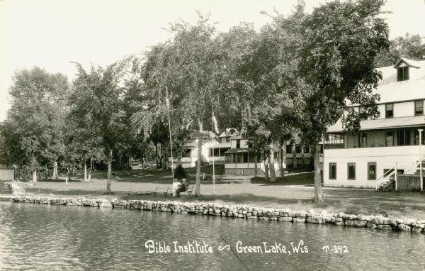 Exterior view across water towards part of the Green Lake Bible Institute, with several other buildings. Two men are sitting on a bench near the lake shore and two flagpoles. Caption reads: "Bible Institute — Green Lake, Wis."