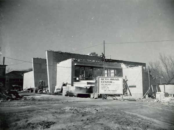 Exterior view of the Beth Israel Center synagogue during construction. A worker is standing on the roof of the building, and construction materials are piled around the base of the building. A large sign in front lists the builders associated with the project: Vogel Brothers Building Co., General Contractor; H. Toussaint, Heating; H. Golden & Son, Plumbing; Cirves Elec. Co., Electrical; John J. Flad & Associates, Architects and Engineers.