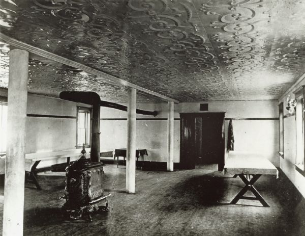 Interior of a dining room, with a wood stove and tables. Caption reads: Pigeon Falls, Wis. 1913. Parish house dining room of Pigeon Creek Norwegian Evangelical Lutheran Church.