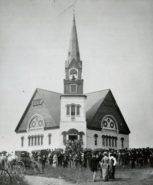 Exterior view of a church, with the congregation entering. Some horse-drawn buggies are on the left. Photograph caption reads: "S.O. Stevens, Stoughton, photog. 1st East Koshkonong Church (upper church). 