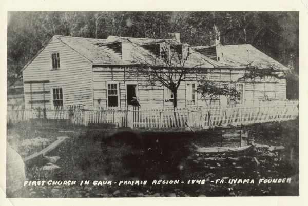A wooden building enclosed by a fence. Caption on rear of photograph reads: First Church In Sauk-Prairie-Roxbury Region - 1846. Reproduction of an early photograph of the combined church and dwelling erected by Father Inama midway between Sauk City and Roxbury about the year 1846 or later. Father Inama can be seen standing just beyond the fence, and in the immediate foreground are the pebbles fringing what is now called Madison Brook, which springs from Crane Lake near Roxbury, and empties into the Wisconsin River about two miles south of Sauk City. The cellar ruins of this building may still be seen about a half mile back from the point where the brook flows beneath Highway 12 between Sauk City and Roxbury. This photograph is of the first church of any kind erected in this region. Reproduction by Arnold Photo Service, Sauk City, Wis.