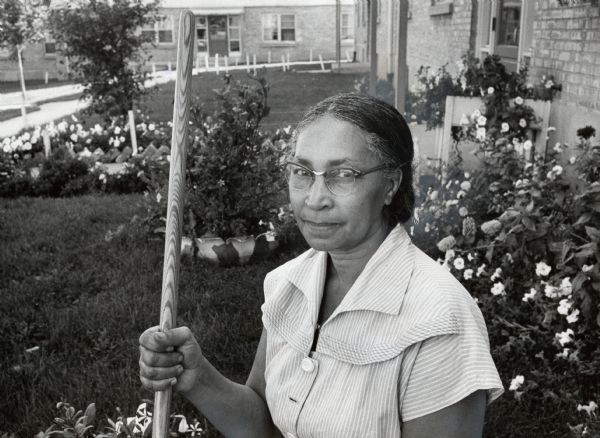 A woman is holding the handle of an unidentified gardening tool and posing in her yard. Caption reads: "Winning one of six plaques for yard beautification at the Hillside Terrace housing project last week was Mrs. Olivia Townsend, 1441 N. 8th St. Two garden clubs, 14 buildings and 121 individuals were honored by the housing authority."