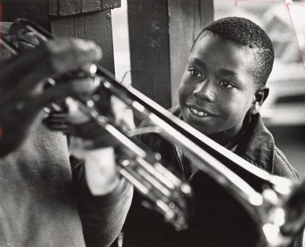 A young man is smiling and looking at a jazz trumpeter, of whom only his hands and trumpet can be seen. Caption reads: "Morris Gray, 11, of 2801 N. 11th St., seemed enthralled by Wetzel's jazz trumpeting."