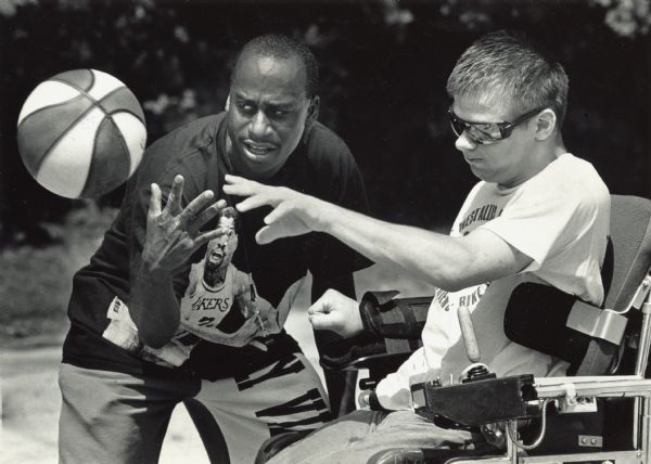 A man is helping another man, who is in a wheelchair, to pass a basketball. Caption reads: "HELPING HAND — Greg Birmingham, coordinator of the Easter Seal Athletic Olympics last Sunday, gave Mark Hubbard a hand in shooting a basketball during a shootout event. The Olympics were held at Holler Park, 5151 S. 6th St."
