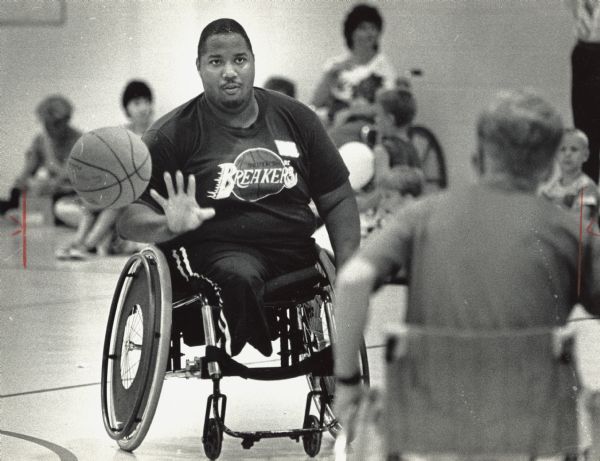 A man in a wheelchair is passing a basketball to someone in the foreground. Caption reads: "The Waukesha Park and Recreation Department, in cooperation with the special summer recreation program, sponsored a disability awareness day Monday to bring together children in the regular playground program with disabled children from the special summer recreation program. Ricky Chones (left) of the South Shore Breakers basketball team passed to teammate Rick Benavides as part of a demonstrations [sic] of the way wheelchairs can be used in athletics."