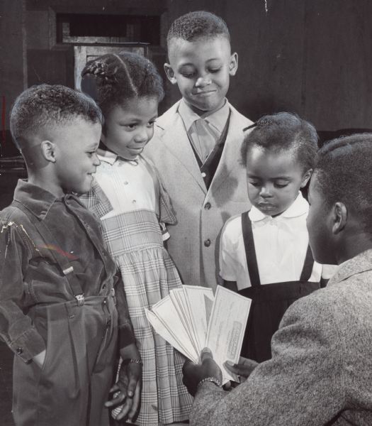 Four children smile at a young man holding out checks. Caption reads: The year's savings of members of the Neighborhood Saving and Recreation organization were distributed at a Christmas party of the children's group Saturday at Antioch Baptist church, 1653 N. 5th st. Melvin H. Jackson (right), 14, of 1746 N. Palmer St., president of the group, gave checks to (from left) John Reed, 4; Hattie Reed, 5, and Elish Reed, 6, of 506 W. Chambers st., and Dorothy Campbell, 4, of 2529 N. 8th St. Totals varied from $1.50 to $13.60.