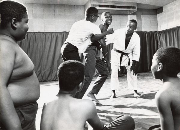 A judo instructor is coaching two young men, while three more young men are looking on. Caption reads: "Instructor Marshall Bullock, 2602 N. 19th st., worked with a group of young wrestling enthusiasts in the North Central YMCA, 2200 N. 12th St." 
Additional notation on the photograph identifies people: Judo class instructor — Marshall Bullock, 2602 N. 19th.
L/R on mat (kneeling) — Larry Townsell, 2371 N. 14th; James Cobbs 325 E. Center; Calvin Morris, 2430 N. 11th
L/R in "combat" — Ealia Beauchamp (white shirt), 2357 N. 6th; Kenneth Kingsby, 2421 N. 6th.