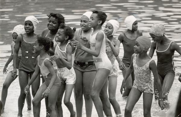 Several girls in swimsuits are standing in shallow water. Caption reads: "IT WAS a brisk day when inner core children visited West Bend park. But the children, accompanied by the Rev. Gene Afseth of Hephatha Lutheran church, 1720 W. Locust St., enjoyed every minute of the visit. A group of girls (left) braved the water and moved about to keep warm."
