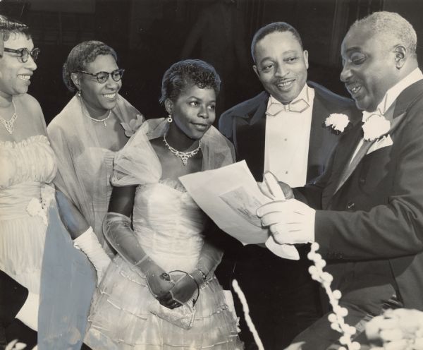 A man is reading from a piece of paper, while a man and three women are looking on. Caption reads: "The first debutante to arrive at the 21st annual ball of the Cosmopolitan Club 25 Saturday night at Prince Hall Masonic temple, 1218 W. North Av., was Miss Sara Lee Blathers, 768 W. Reservoir Av. She was greeted by Mrs. Roeshell Steward (left) 2915 N. 11th St.; Raymond Renfro (right), 1537 N. 9th St., social secretary of the club and program chairman, and Roeshell Steward, president of the club.