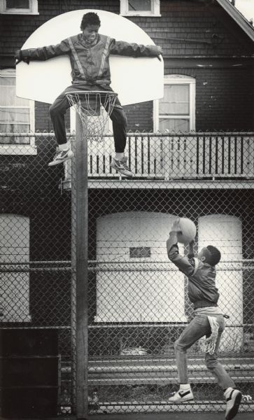 One boy is sitting up against the backboard of a basketball hoop and looking down, while another boy is preparing to shoot a basket. Caption reads: "Terrell Henderson had a bird's-eye view from atop the basketball rim as his friend, Damon Simpson, took a shot Sunday at a basketball court at N. 33rd St. and W. North Ave."