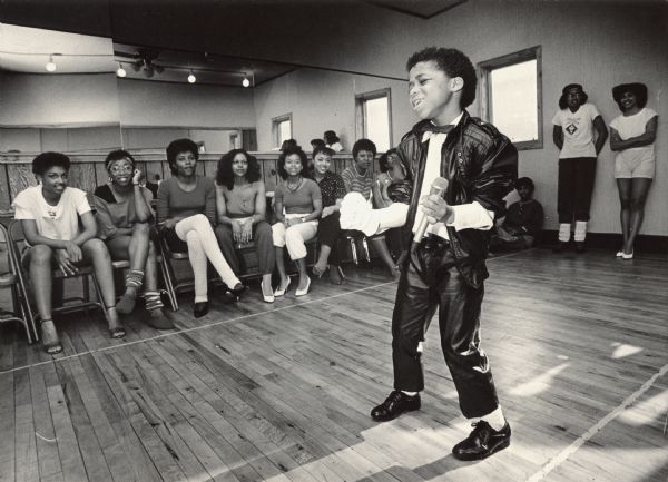 A boy dressed as Michael Jackson is singing as a group of young people are watching in the background. Caption reads: "Six-year-old Jamal Stevenson imitates singer Michael Jackson during pageant rehearsals."
