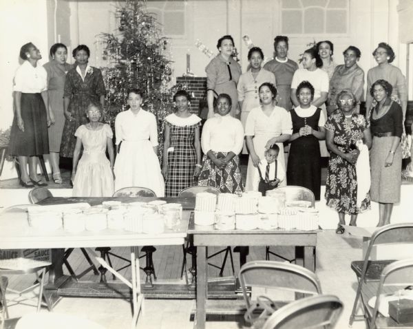 Several women and children are standing and posing, with a Christmas tree behind them, and tables in front of them that have gifts on them. Caption reads: "Milwaukee, Wis. 1957. "Mrs. Addie Taylor — Home Nursing Neighbors — Christmas Party - Urban League Auditorium."