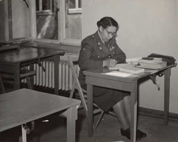 A woman in military uniform is sitting at a desk writing in a notepad. Caption on back reads: "Cpl Daisy Berry, Milwaukee, Wisc., a member of the 7871st Training and Education Group WAC [Women's Army Corps] Detachment at the Kitzingen Training Center is the only WAC in the European Command who participates in the on-duty education program. (PIO PHOTO BY JOHNSON)."