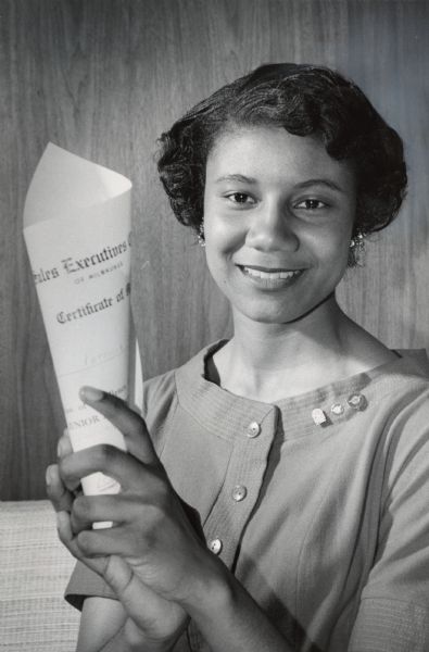 A young woman is smiling and holding up a certificate. Caption reads: "The top salesmen in the Junior Achievement sales contest showed her certificate proudly at an awards ceremony Thursday night at the Milwaukee Athletic club. She is Patricia Williams, 18, of 223 N. 32nd St., who was also awarded a $100 prize. She topped 1,200 Milwaukee Junior Achievers."