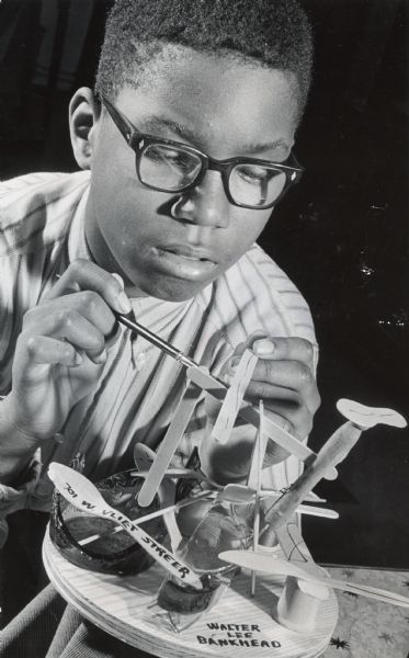 A boy is working on an art project. Caption reads: "Art projects are popular this month at St. Benedict's school, 1004 W. State St. The children have had no help from teachers in selecting materials or ideas. Walter Bankhead, 12, of 701 W. Vliet St., used wooden spoons and tongue depressors in his project."