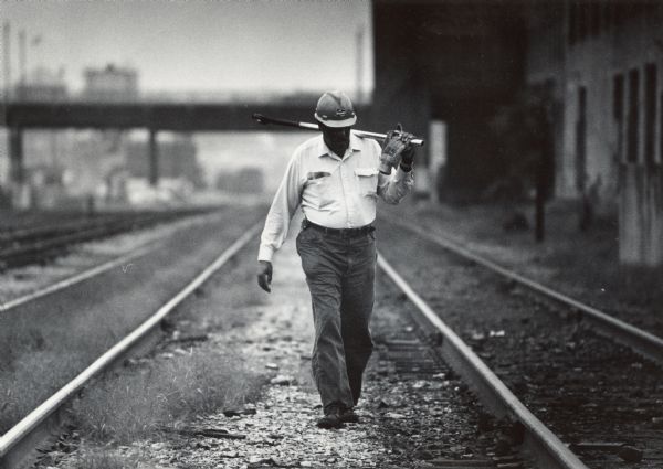 A man wearing a hardhat and carrying a tool is walking along railroad tracks. Caption reads: "A railroad worker walked along a line in the Menomonee Valley near Muskego Ave."