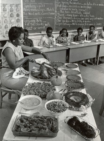 A woman is carving a turkey, which is on a table full of many dishes. Several children are sitting and looking on from behind their desks. Caption reads: "AND ALL THE FIXINGS — Third grade children at the Palmer Street school waited anxiously for their teacher to carve a 25 pound bird Tuesday morning. Mrs. Eva Andrews, 2540 N. 2nd St., brought a complete Thanksgiving dinner to class. Thirty-one children feasted on turkey, salads, dressing and pumpkin pie."