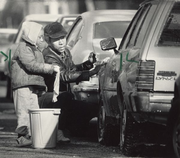 A man and his son washing their car together. Caption reads: "Victor Driver, 4, helped his stepfather, Thomas Holder, wash his car Sunday on N. 13th St. as relatively warm weather enabled area residents to perform outdoor chores in comfort."