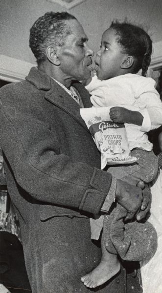 A man is holding a young girl in his arms. She is holding a bag of Geiser's potato chips. Caption reads: "Poverty faces this father's six daughters, 3 to 12 years old, as he approaches the age of 65. Now, at 62, he earns about $95 to $100 a week as a tannery worker."