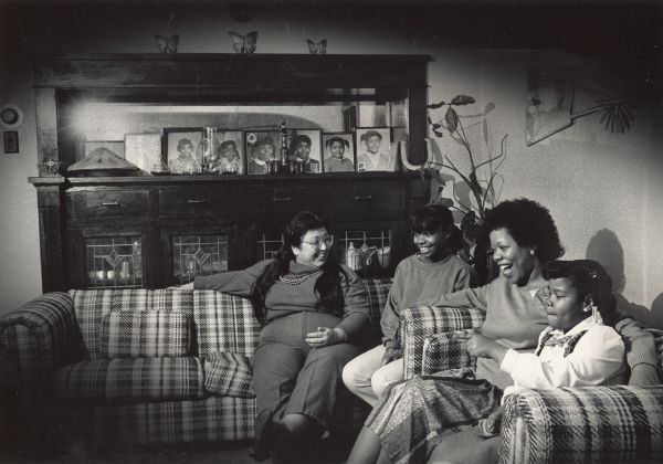 Two adult women and two girls are sitting on sofas and smiling. On a shelf on a mirrored cabinet behind the sofas are several framed photographs of girls. Caption reads: "Eleanor Walker (second from right) and her daughters, Erica (right), 7, and Joy, 12, relaxed with their friend Diana Lualhati (left), after a Thanksgiving Day meal."