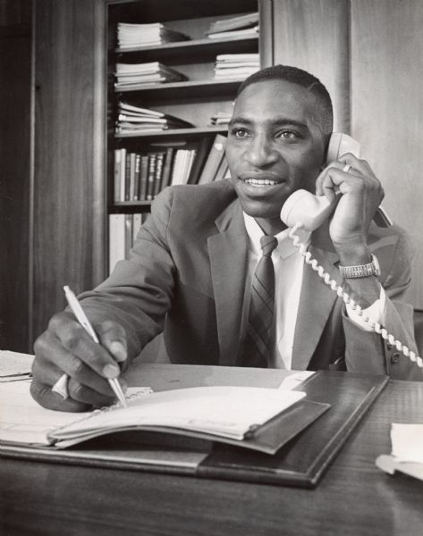 View across desk towards a man talking on a telephone and writing in a notebook. Caption reads: "Adam Davis, who is an insurance salesman, represents the growing Negro middle class."