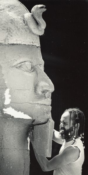 A man is grimacing while working on what appears to be an Egyptian stone sculpture. Caption reads: "Tejumola F. Ologboni attached the beard to a figure on the float representing Afro Fest in the City of Festivals Parade to be held June 20. The float is one of several under construction at 301 E. Walnut St."
