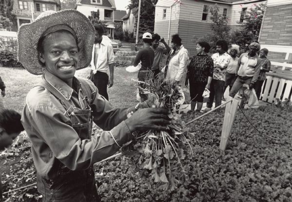A man is smiling while holding up a handful of greens from a garden. In the background, several people are standing in a lined. Caption reads: "Milwaukee resident Eddie Patterson held up a handful of greens that he planted at his aunt's home at 2406-A N. 7th St. this spring. Patterson was giving them away Friday to anyone who came by. By the end of the day, all of his greens, turnips, and spinach were gone. He said he gave them away because people needed the food, and he thought that the drought would make vegetables expensive. Patterson planted his garden about a month ago and vigorously watered and fertilized it. He plans to plant greens again next week and, when they're grown, give those away too."