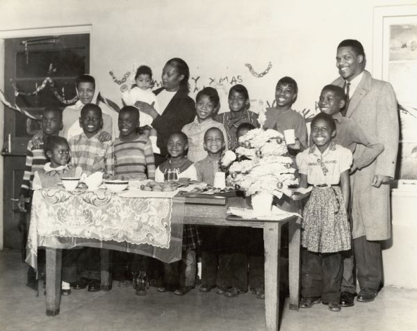 A group of smiling children posing with three adults. They are standing behind a table covered with baked goods, and Christmas decorations are on the wall behind them, and on the table in front of them. Caption reads: "Norman Johnson — Hillside Block Unit — Midget Unit Christmas Party."