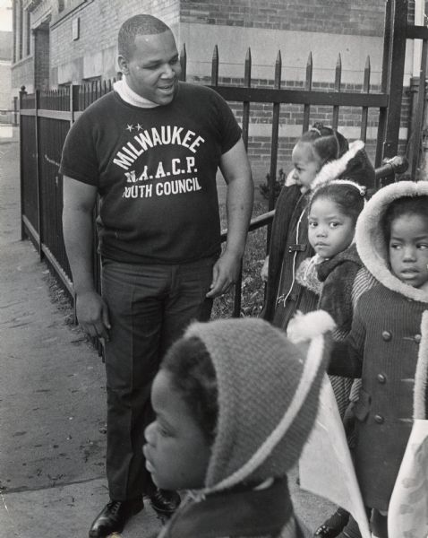 A young man in a Milwaukee N.A.A.C.P. Youth Council t-shirt is standing and talking to a group of children. Caption reads: "Richard E. Green, 22, of 3233 N. 10th St., talked with youngsters from La Follette elementary school, 3239 N. 9th St., as they left for home Tuesday afternoon. Green, a commando with the National Association for the Advancement of Colored People youth council, worked for the school as a 'building attendant' Tuesday and Wednesday when schools have parent-teacher conferences."