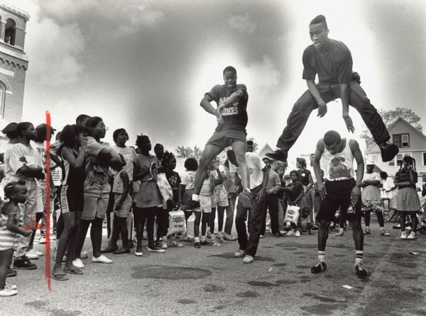 Four young men are performing a dance routine outdoors (two are leaping in the air), while a group of people is watching them. Caption reads: "The dance group Creativity, including Shawn Fisher, Ian Hayes, Johnny King, and Reginald Yarbrough, performs a dance routine at a cookout at the Social Development Commission's Westside Neighborhood Center, 2449 N. 36th St. Friday's event featured food, musical entertainment, children's games and informational presentations by social service agencies."