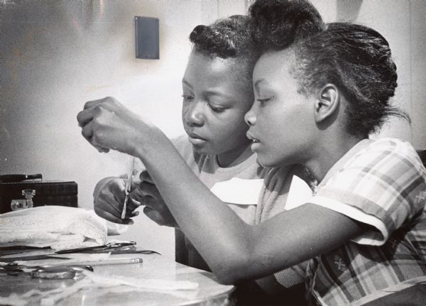 Two girls are focusing on cutting a piece of lace. Caption reads: "Junior homemakers are intent with their work, making Christmas stockings. Delores Taylor, 9, of 817 W. Vliet St., and Rosetta Walters, 10, of 810 W. Vliet St., were among the girls who attended a sewing session at the Hillside public housing development."