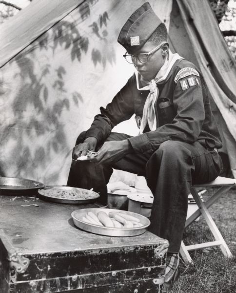 A boy in a Boy Scouts uniform for Milwaukee County Council 406 is sitting on a camp stool and peeling carrots. Caption reads: "Peeling carrots was unexciting business for Hebert Renfro, 16, of 2629 N. 7th Lane, with so many other interesting things going on near him."