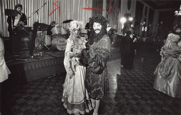 Two people in costume posing on the dance floor. A few other dancers and band members are in the background. Caption reads: "Masked Ball — At the second annual Charity Masquerade Ball of the Syrena Polish Folk Dance Ensemble Saturday night, co-chairs Alicja Newell and Neil Dziadulewicz took a turn on the floor in the Empire Room of the Marc Plaza Hotel. Proceeds were earmarked for the Polish Relief Fund."