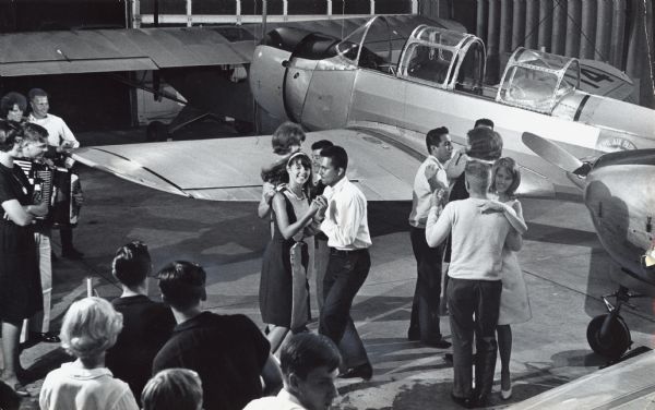 Elevated view of four couples dancing near airplanes, while several other people look on. One person is playing the accordion. Caption reads: "They moved the planes aside at Timmerman field Tuesday night for a Civil Air Patrol party. The dance and ice cream social honored five CAP cadets from El Salvador who are visiting the Milwaukee area this week as part of an international cadet exchange."