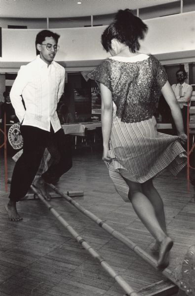 A man and woman performing a dance over two bamboo poles. Caption reads: "Students from around the world brought their customs and culture to Marquette University Wednesday for International Day 1989. Freshman Ed Fernandez and sophmore Bernadette Davantes performed a Filipino dance called tinikiling, named after a bird."