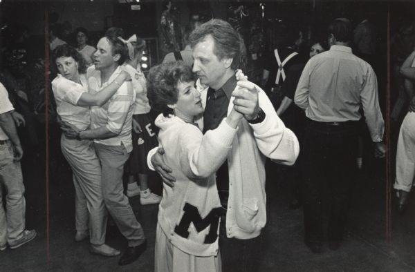 A couple is dancing in the foreground while several people are dancing in the background. The woman is wearing a sweater with an M stitched to it. Caption reads: "Helen and Jim Alherton danced to '50s and '60s music. He is a '63 graduate of Menomonee Falls High School; she's a '63 Messmer grad."