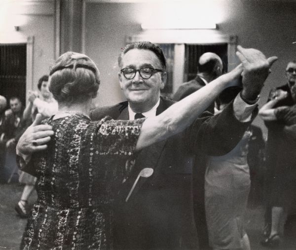 A man and a woman are dancing, with other people dancing in the background. Caption reads: "Charles Denny showed his delight as he waltzed with a guest, Mrs. Minnie Rolin, 2435 N. 10th St."